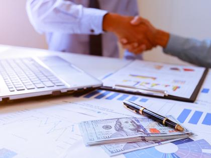 Two people wearing dress shirts shaking hands near a table with an open laptop. Multiple papers and $100 bills are on the table.