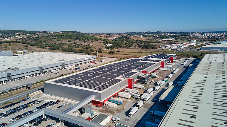 An aerial view of a warehouse, the roof is covered in solar panels, many trucks are entering and exiting the loading dock area