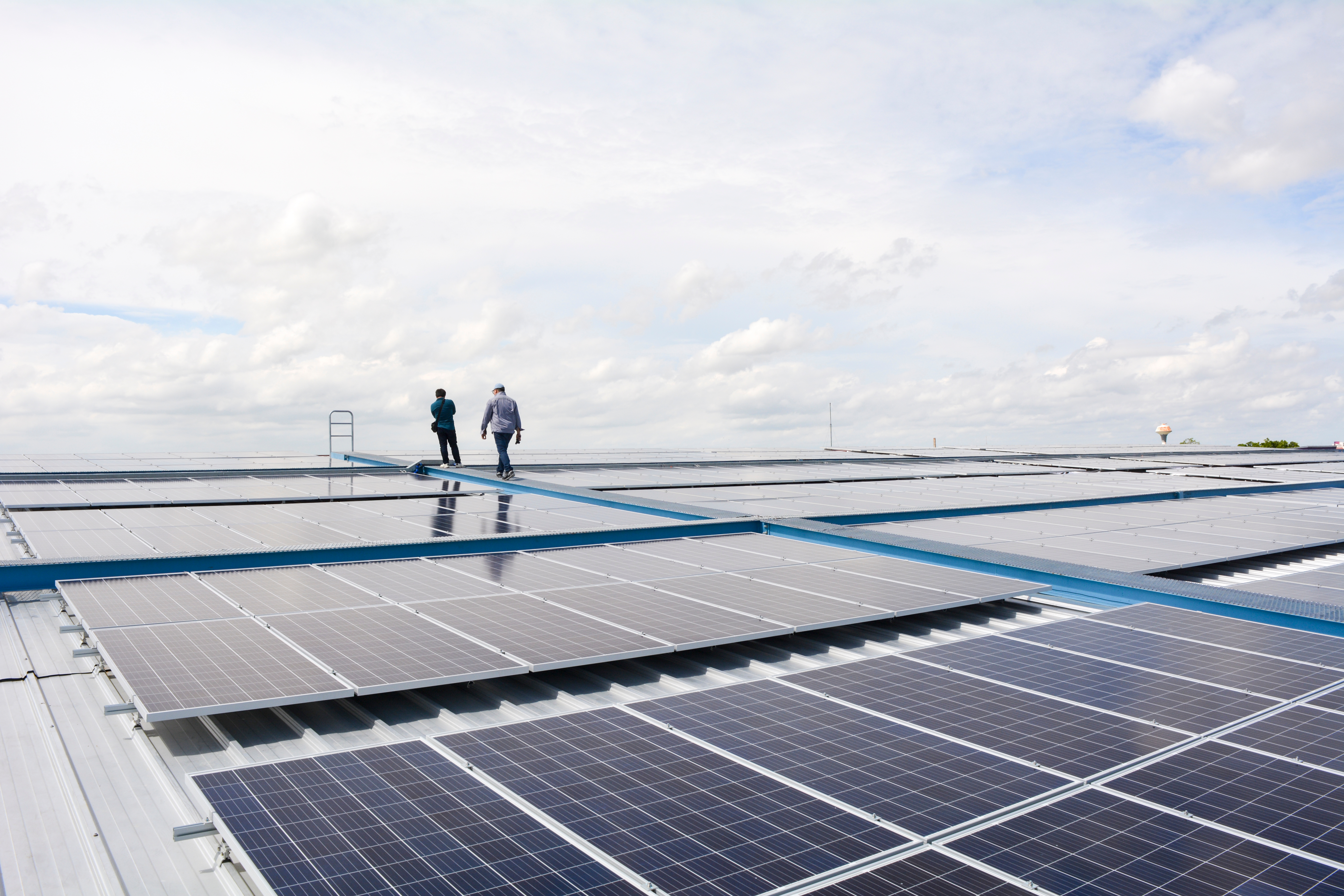Two men standing on the rooftop of a building covered in solar panels on a sunny yet cloud filled day.