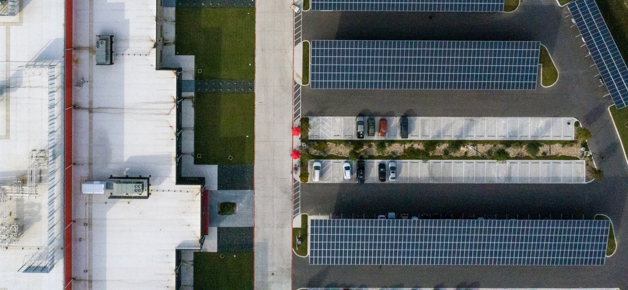 Aerial view of a parking lot with solar panels and a handful of cars, next to a large building with a white roof.