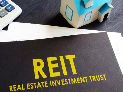 A black folder labeled REIT (Real Estate Investment Trust) in yellow type on a desk with a calculator and a toy house