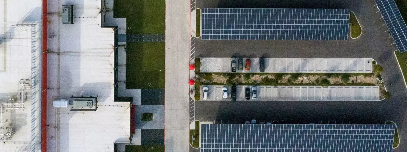 Aerial view of a parking lot with solar panels and a handful of cars, next to a large building with a white roof.